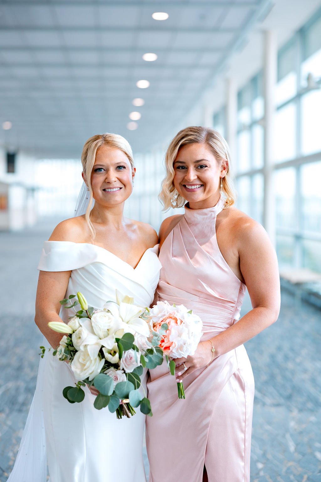 Two women at a wedding, one in a white wedding dress with a veil, and the other in a pink satin dress, both holding bouquets of flowers, standing in a modern, brightly lit indoor space with large windows.