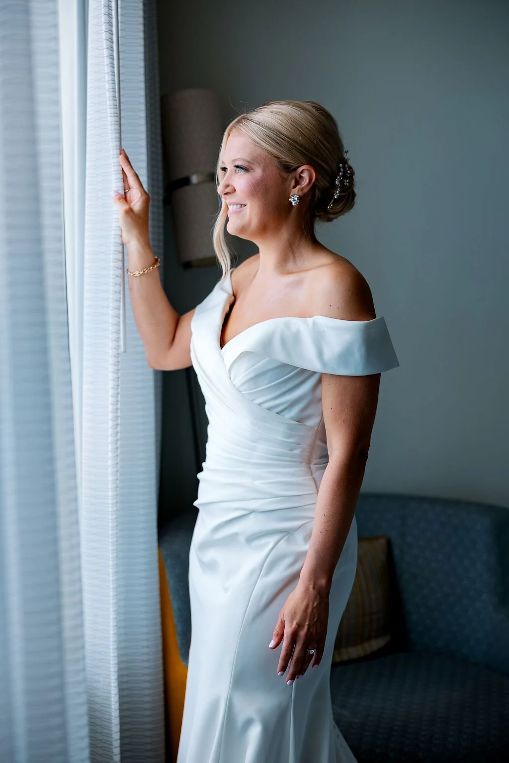 A smiling blonde woman in a white off-shoulder wedding dress looking out a window and holding the curtain.