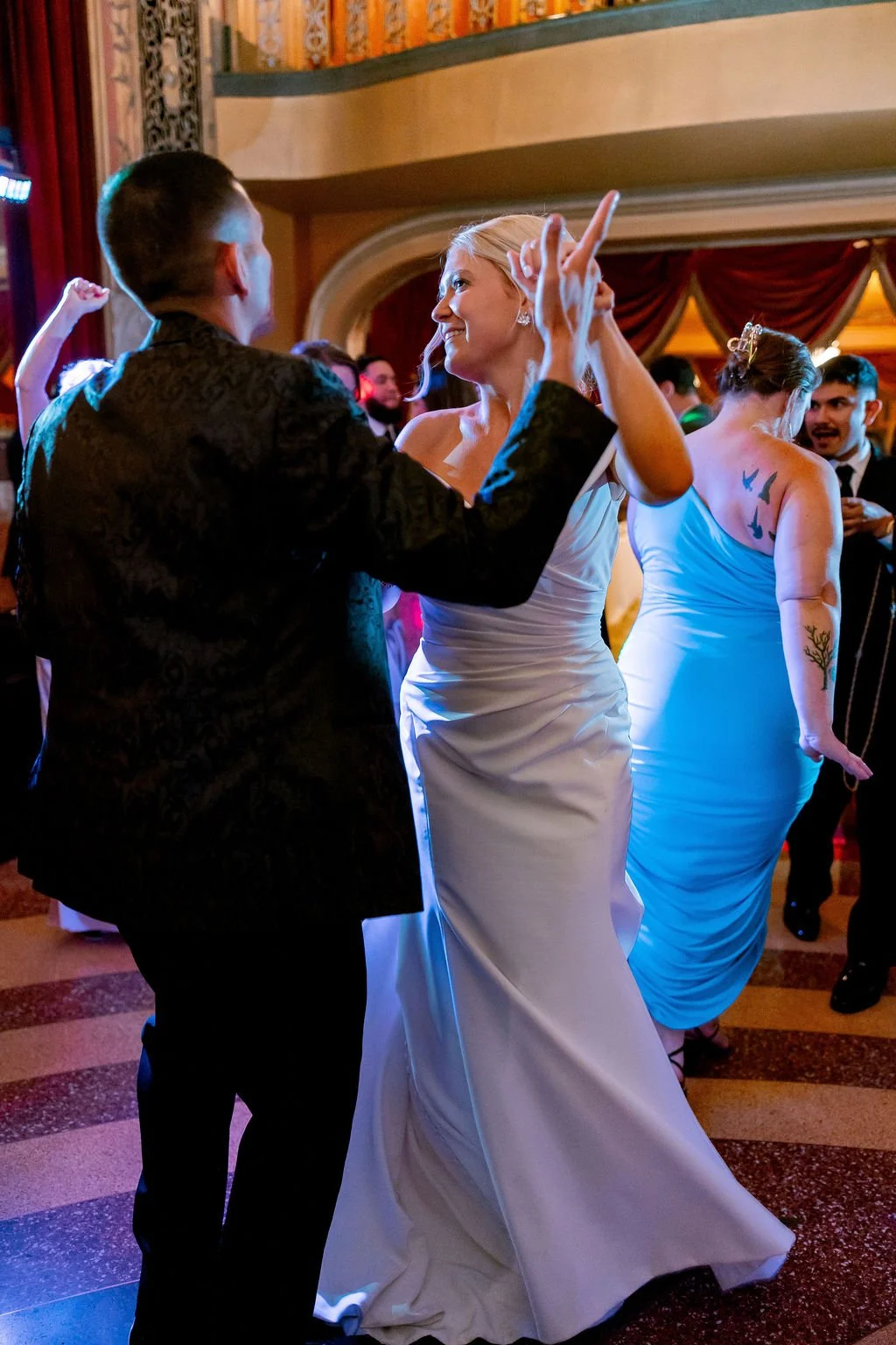 People dancing at a formal event, including women in dresses and men in suits, with colorful lighting and ornate decorations.