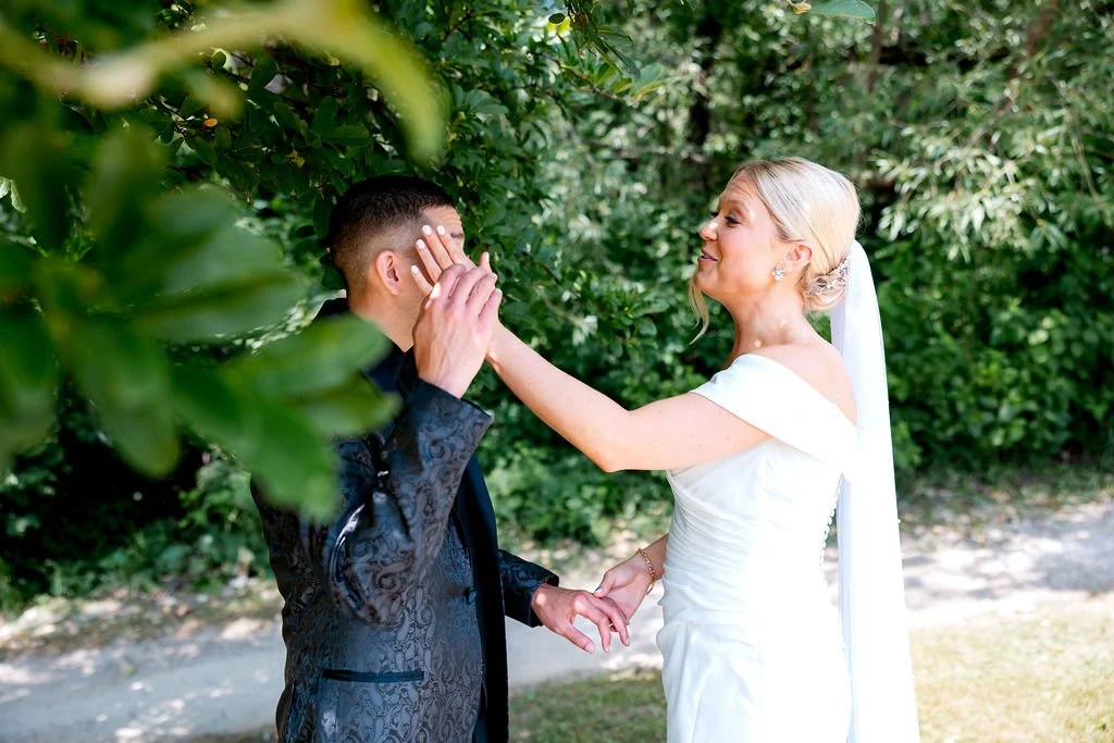 A bride and groom standing outdoors among green trees, with the bride gently touching the groom's face during a wedding ceremony or moment.