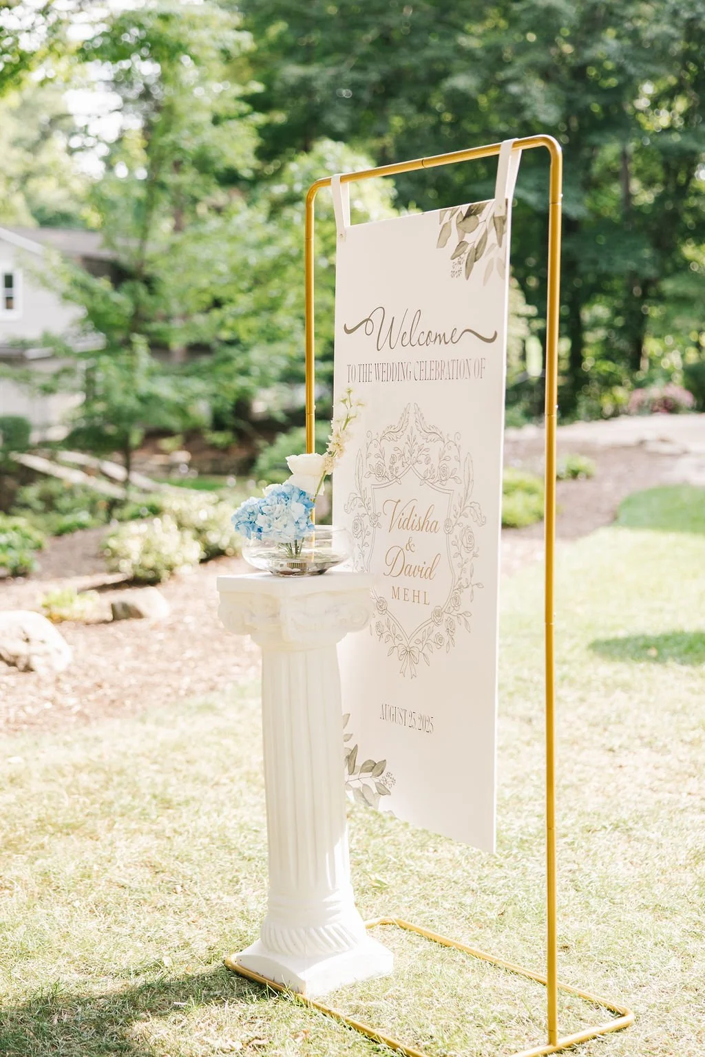 Wedding welcome sign outdoors, decorated with a bouquet of blue and white flowers, standing on a small white pedestal, with green trees in the background.