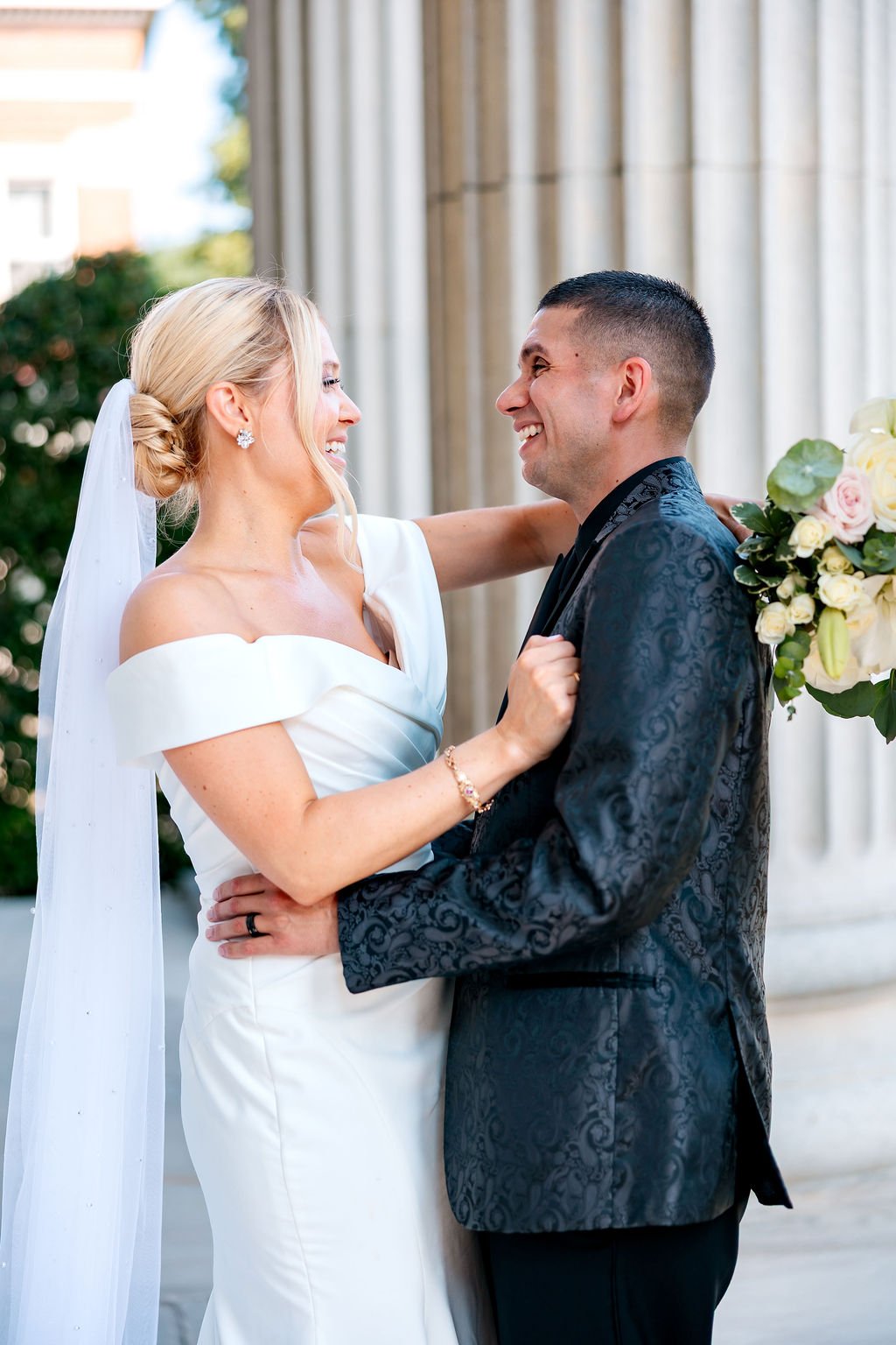 A bride and groom smiling and embracing outdoors during a wedding ceremony, with the bride in a white dress and veil, and the groom in a dark suit holding a bouquet of white and pink roses.