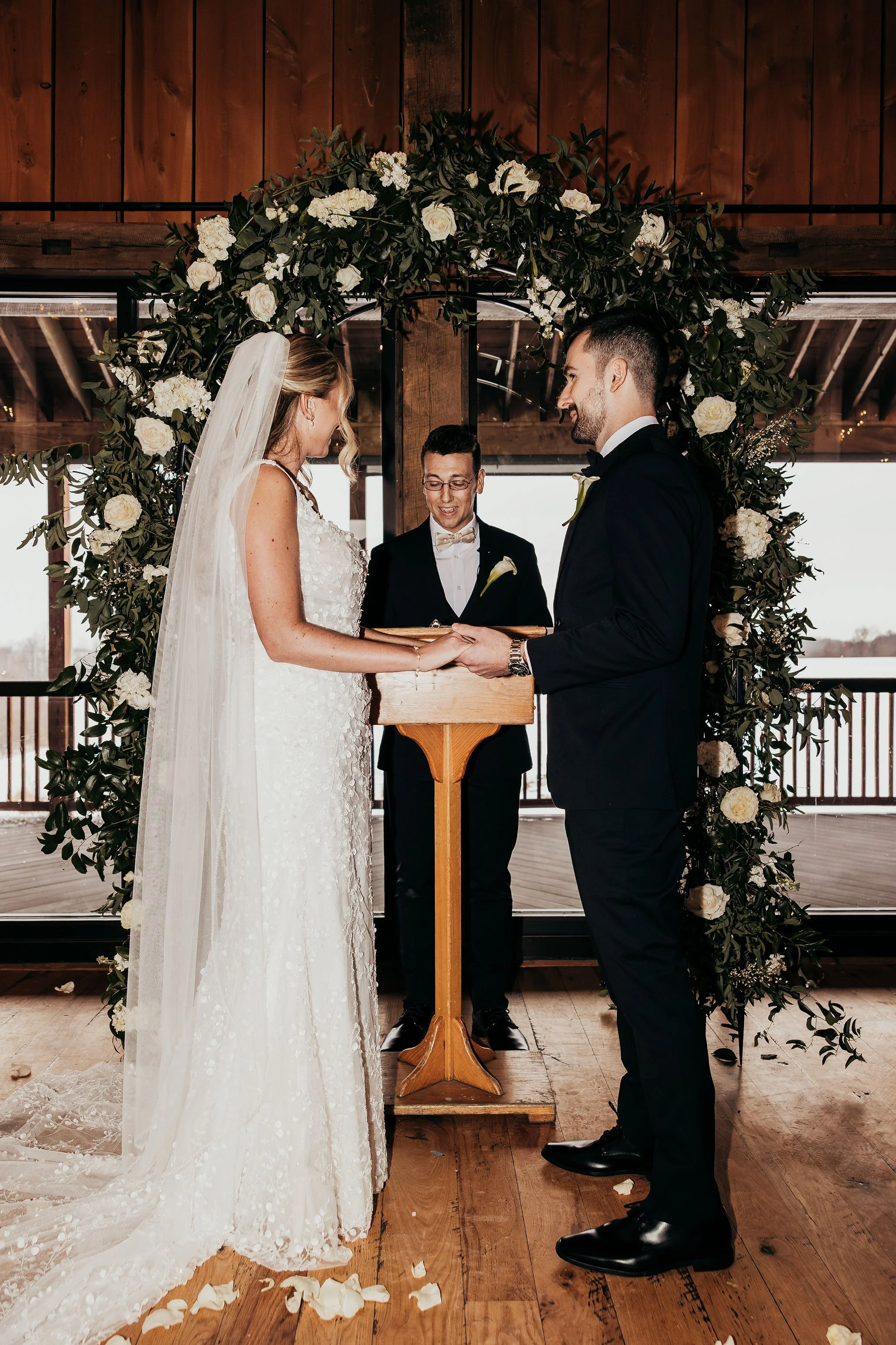 A bride and groom standing under a floral arch during their wedding ceremony, exchanging vows, with a minister officiating, in a wooden venue with windows in the background.
