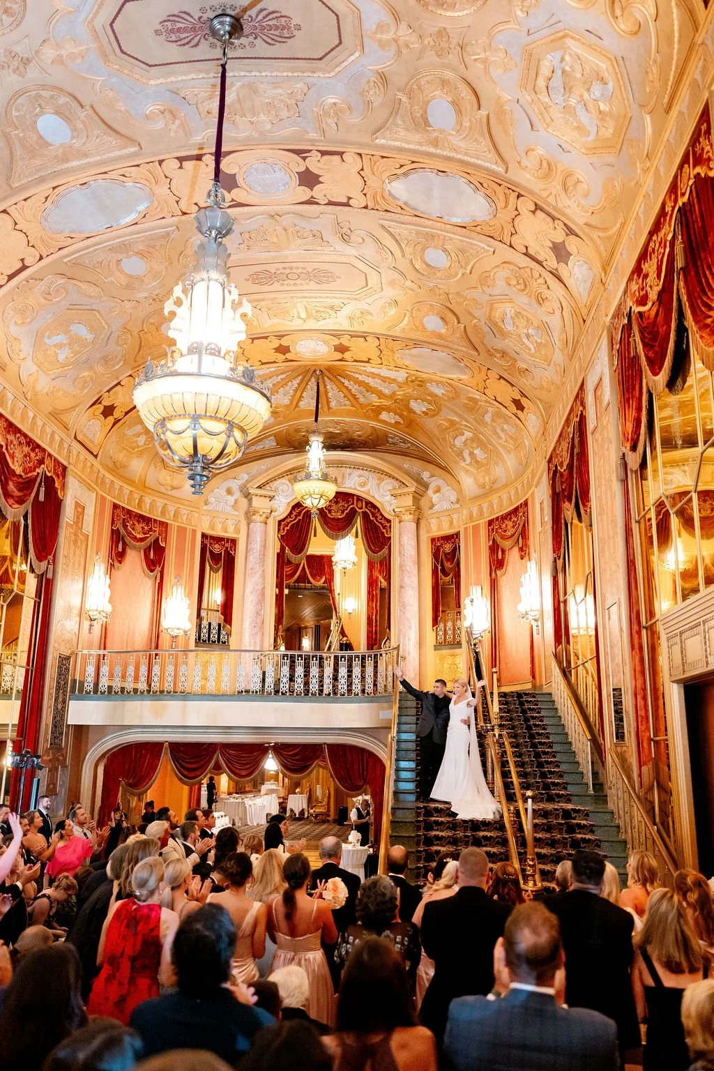 A wedding reception in an opulent hall with a high, decorated ceiling and hanging chandeliers. The bride and groom are on the grand staircase, surrounded by guests watching and applauding.