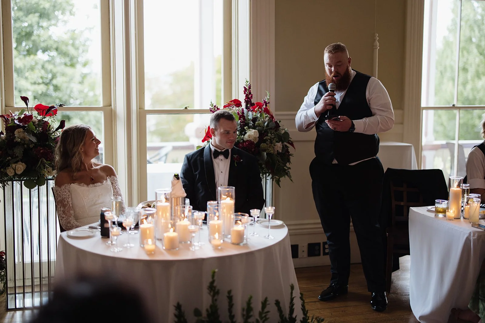 Wedding reception with bride and groom seated at a table decorated with candles, and a man giving a speech while holding a microphone.