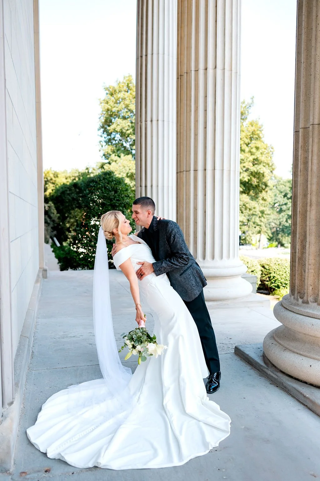 A bride and groom in wedding attire are standing outside between large classical columns, sharing a joyful dance or embrace. The bride is in a white wedding dress with a long train and veil, holding a bouquet of white flowers, while the groom is in a