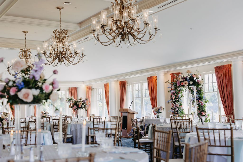 Elegant banquet hall with multiple round tables, floral centerpieces, large windows, orange curtains, chandeliers, and a decorated archway for a wedding ceremony.