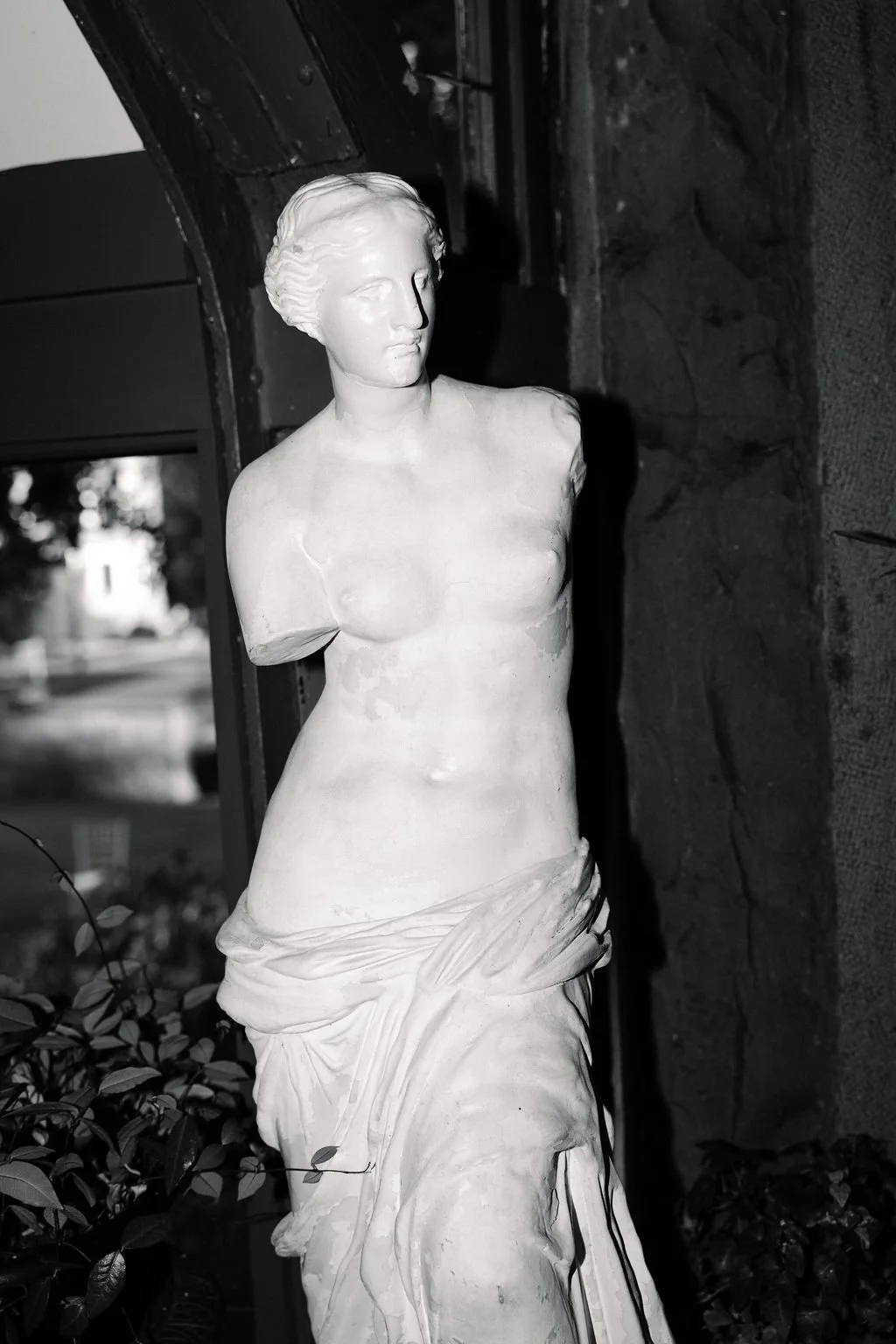 Black and white photo of a classical-style female marble sculpture, missing arms, partially draped in cloth, placed near a window with trees visible outside.