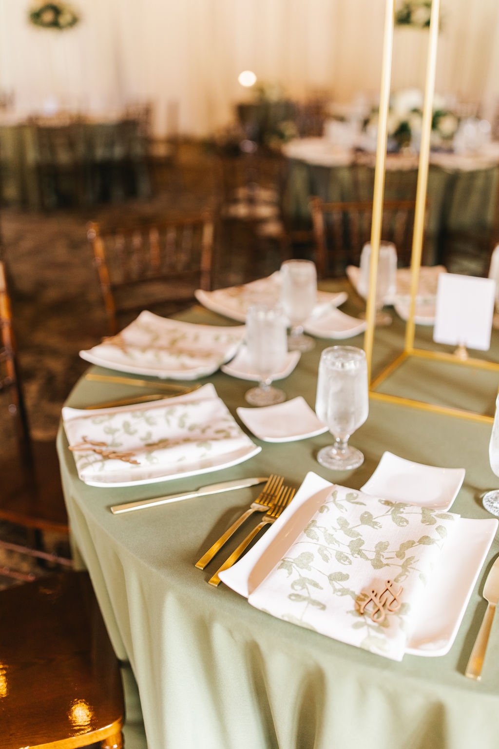 A table set for a formal event with white plates, folded napkins with green leaf patterns, gold utensils, water glasses, and a gold stand with cards or decorations in the center. The background shows more tables and chairs in a decorated venue.