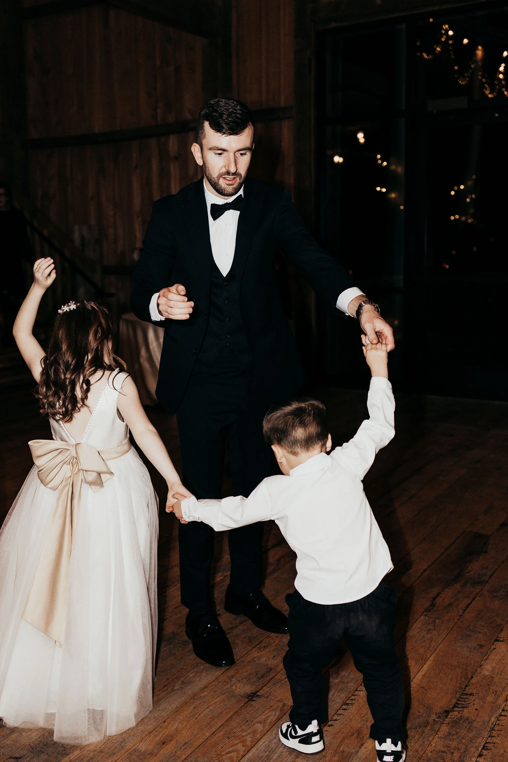 Man in a black tuxedo dancing with children at a party in a dimly lit venue with wooden floors and walls, and string lights in the background.