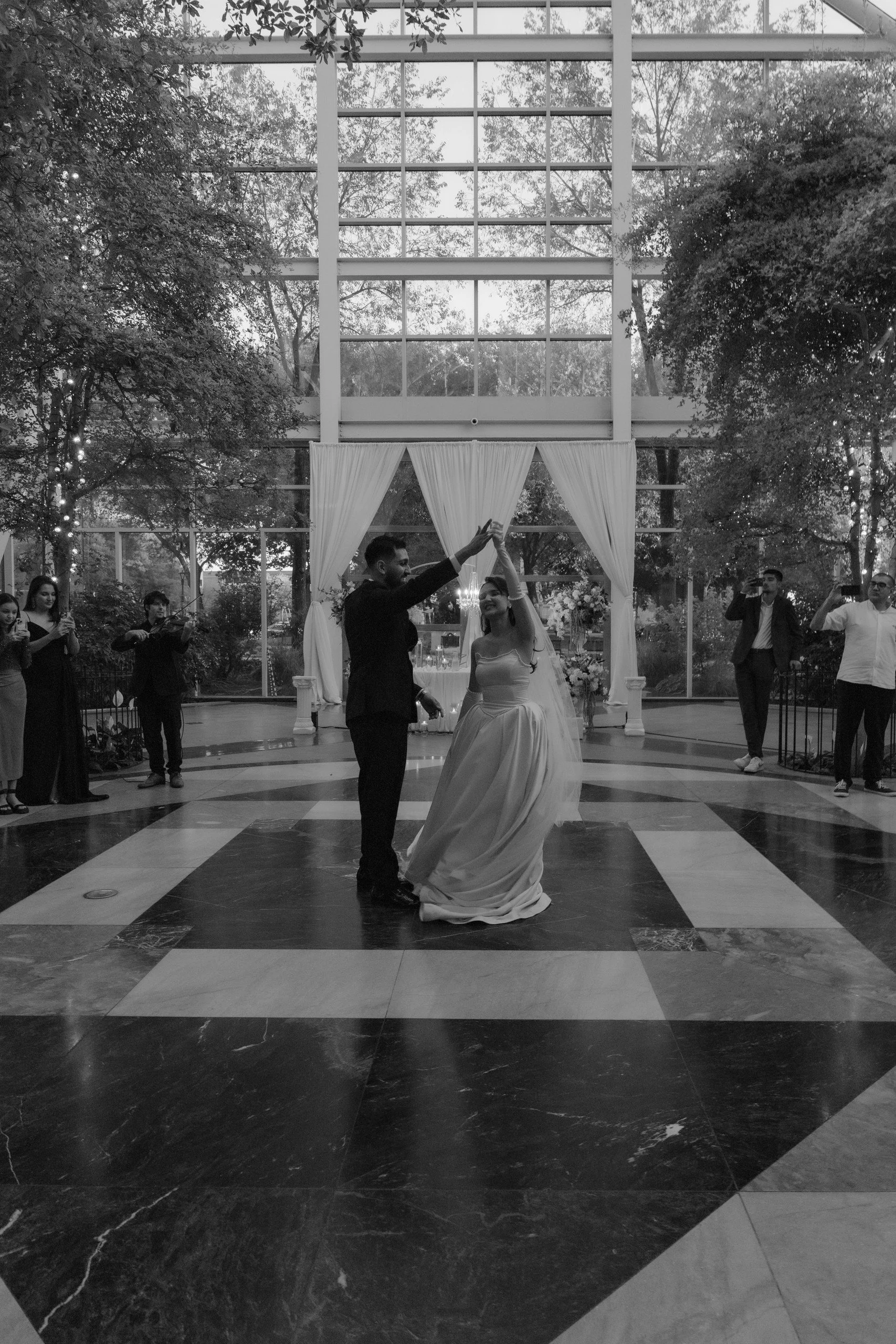 A black and white photo of a bride and groom dancing together in a formal setting, with several guests watching and taking photos. The venue has large windows, trees outside, and decorative curtains.