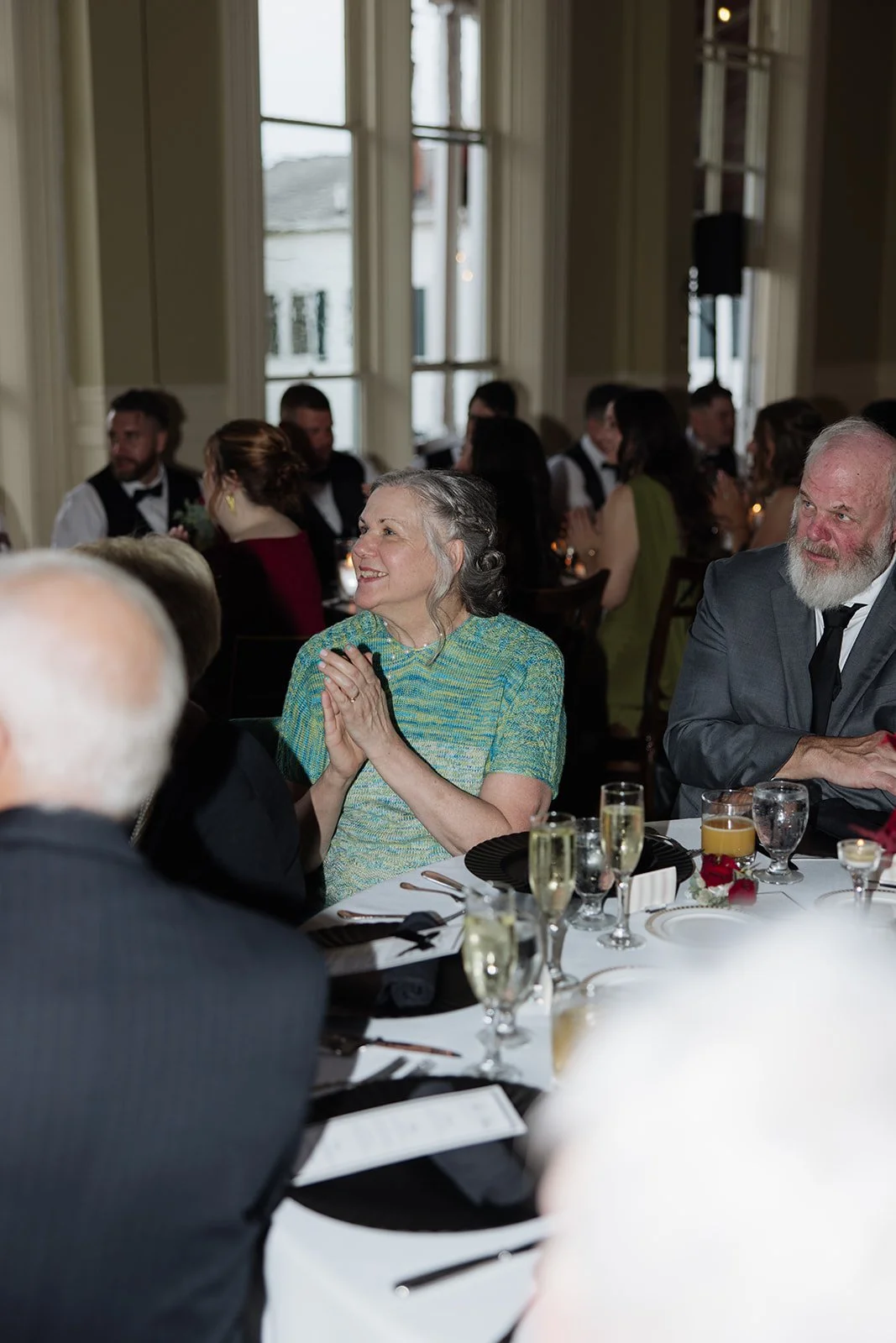 People seated at a formal dinner table, smiling and clapping, with glasses of wine and champagne, in a well-lit room with large windows.