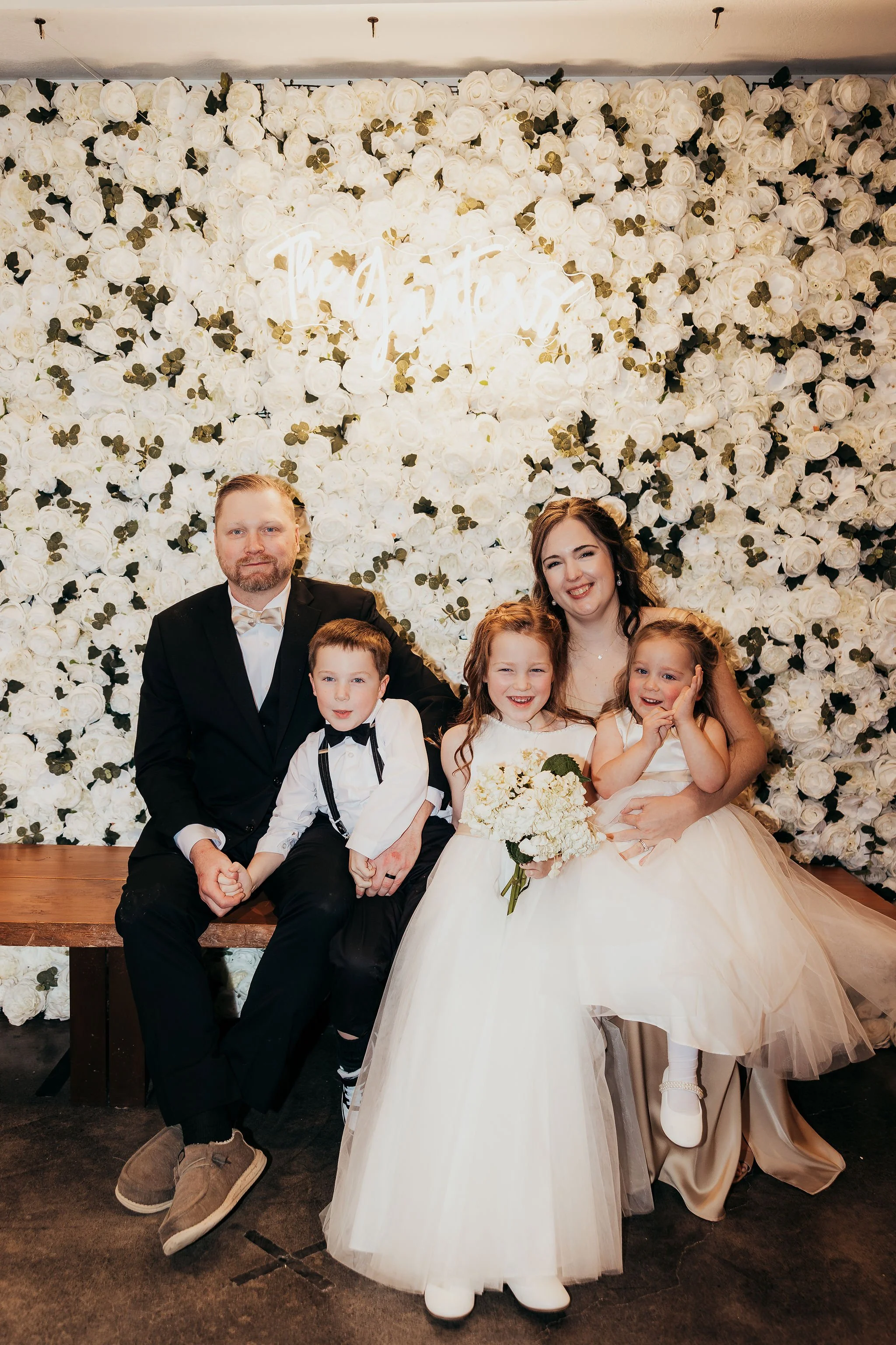 Family portrait at a wedding with a bride and groom, two young boys, and two young girls, sitting on a wooden bench in front of a wall covered with white roses and green leaves, a neon sign that reads 'Together' above them.