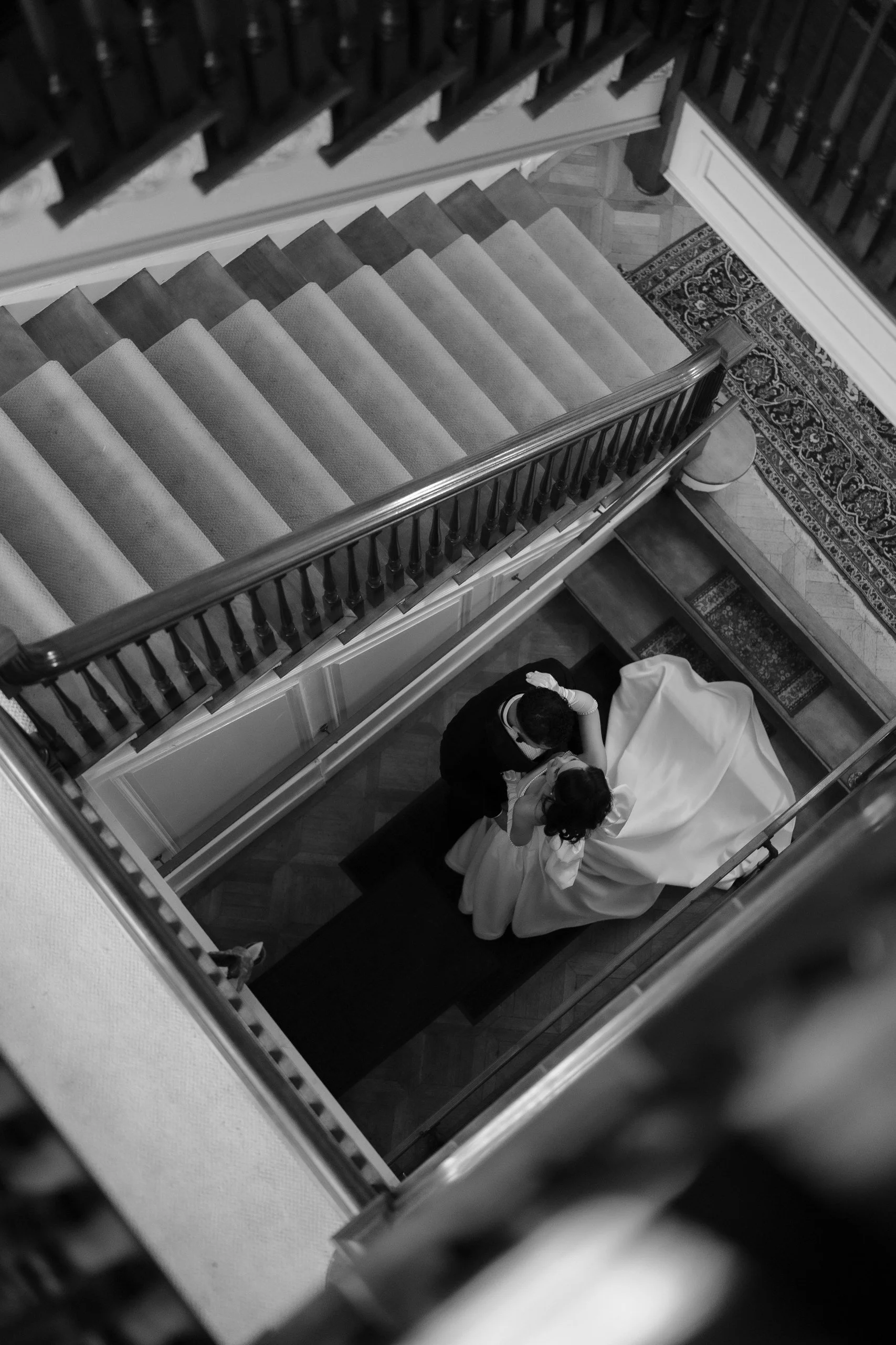 Two people standing on a staircase, apparently preparing for a formal event, with a person in a white gown and another in black, viewed from above.