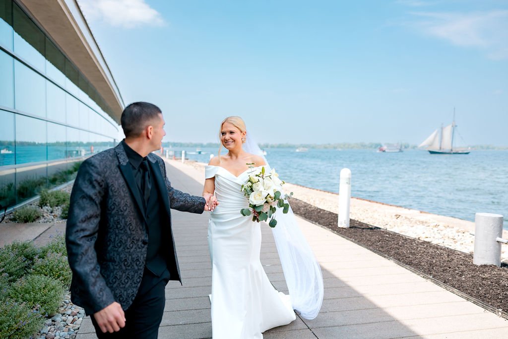 A bride and groom holding hands and smiling as they walk along a waterfront promenade with a sailboat on the water in the background.