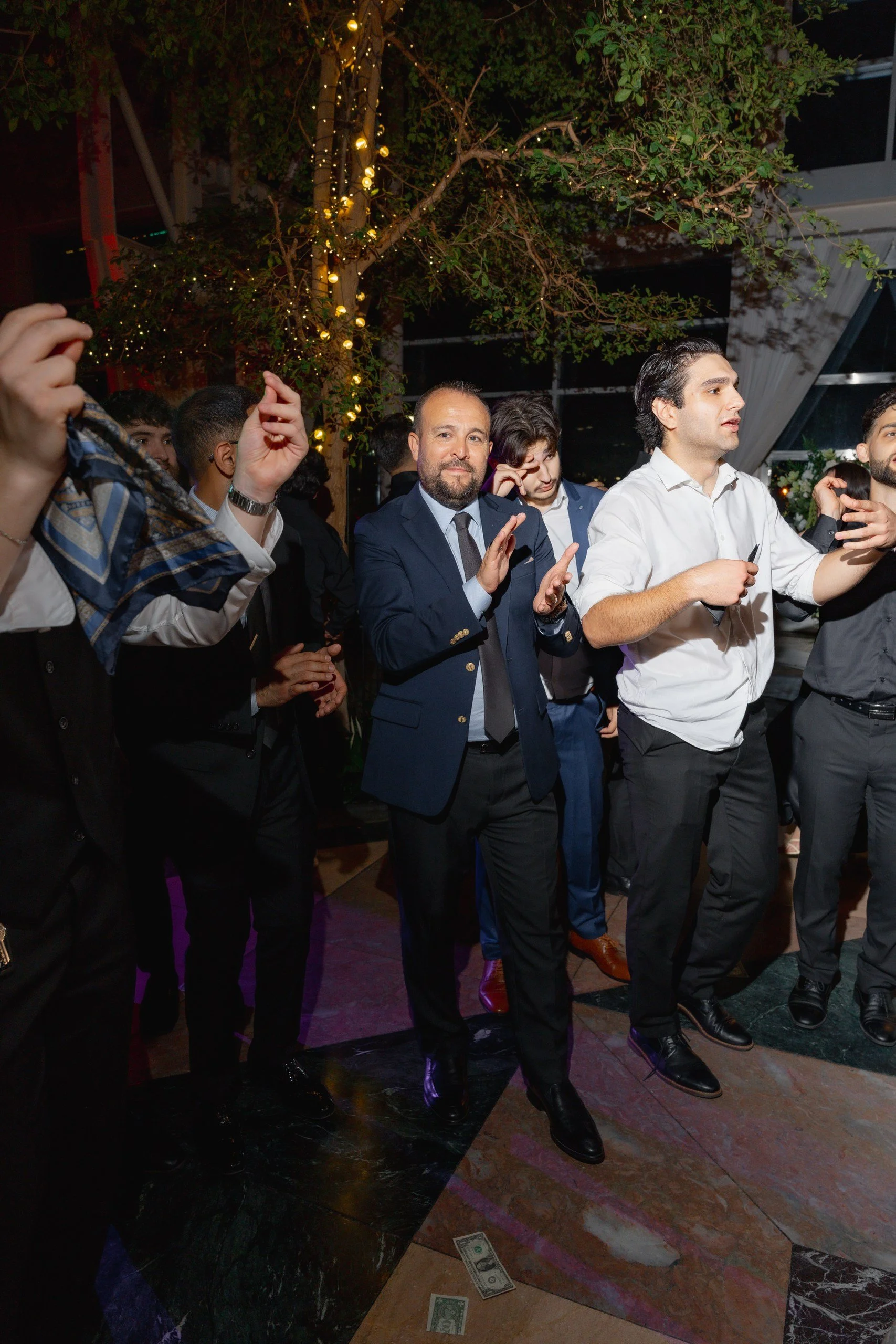 A group of men in formal attire standing together at a social event, with a tree decorated with string lights in the background. US dollar bills are seen on the marble floor in front of them.