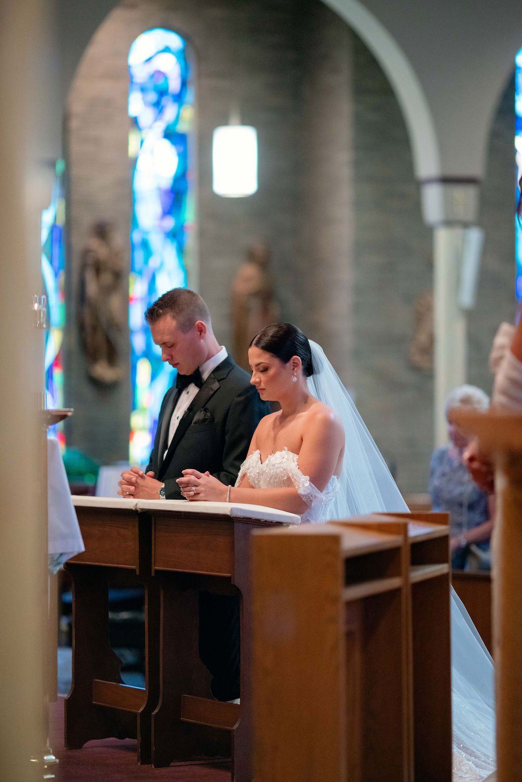 A bride and groom praying during their wedding ceremony in a church with stained glass windows.