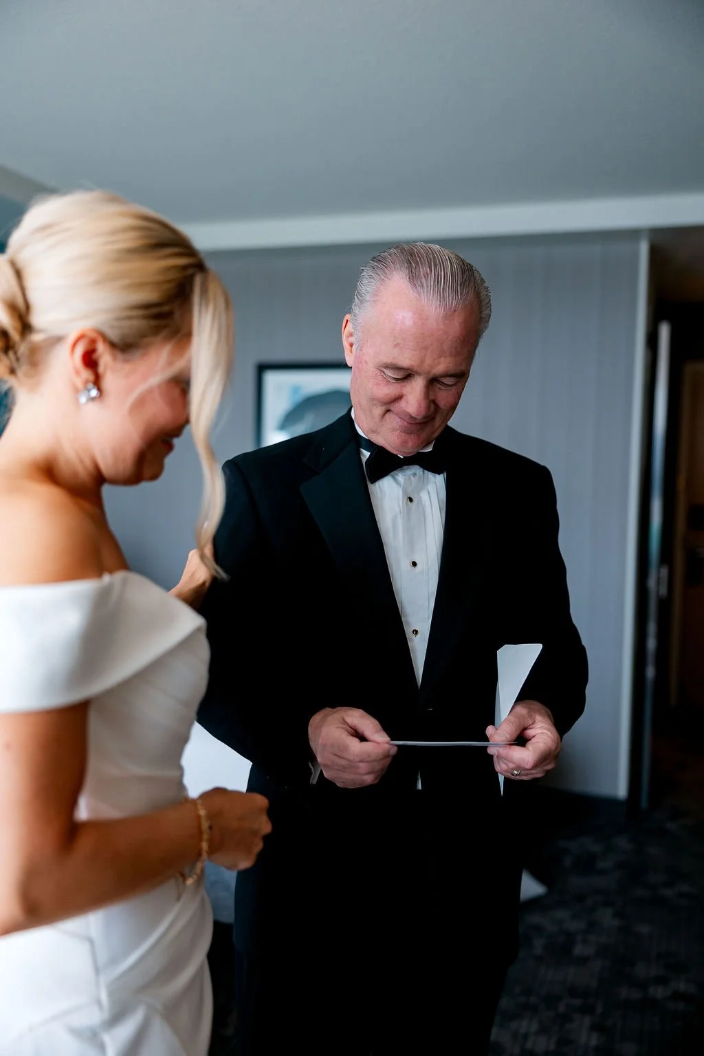 A man in a tuxedo with a bow tie and a woman in a white dress are smiling and looking at a piece of paper, likely during a wedding or formal event.