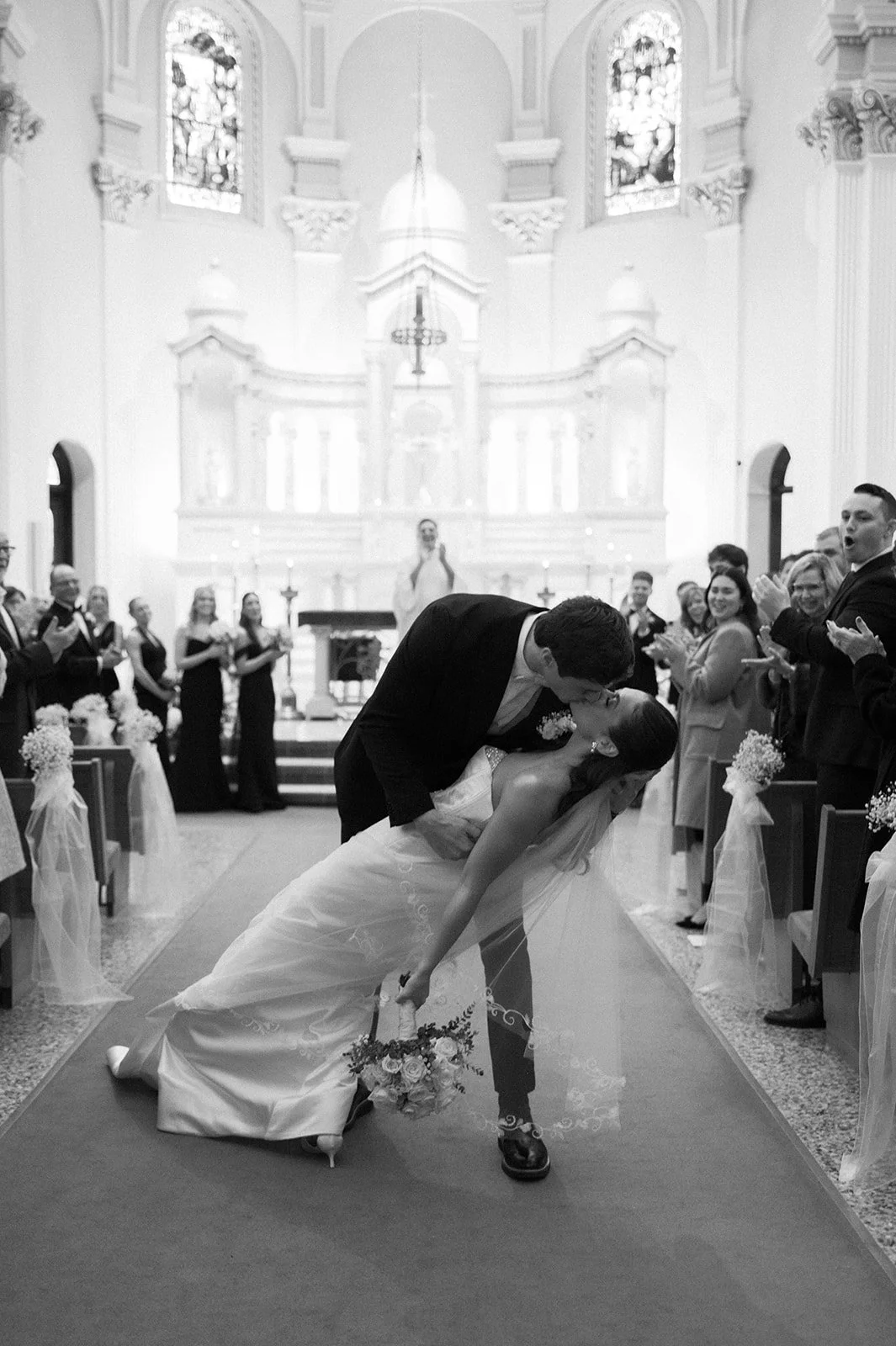 Wedding ceremony in a church with a bride and groom kissing, surrounded by guests clapping and smiling, with floral decorations and stained glass windows in the background.