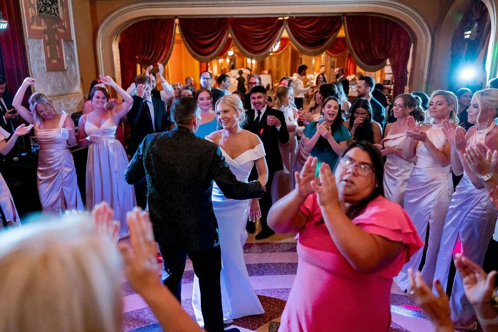 A wedding reception with guests dancing and celebrating inside a ballroom decorated with red curtains and ornate architecture.