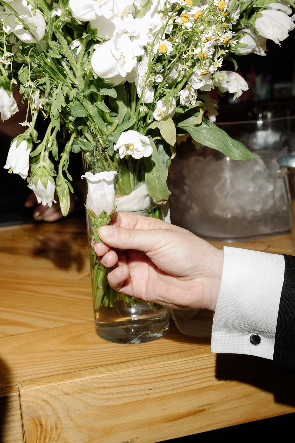 A person holding a white flower bud in front of a bouquet of white flowers in a glass vase on a wooden table.