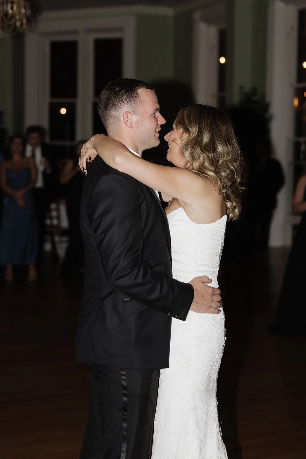 A newlywed couple shares their first dance at a wedding reception, with guests observing in the background.
