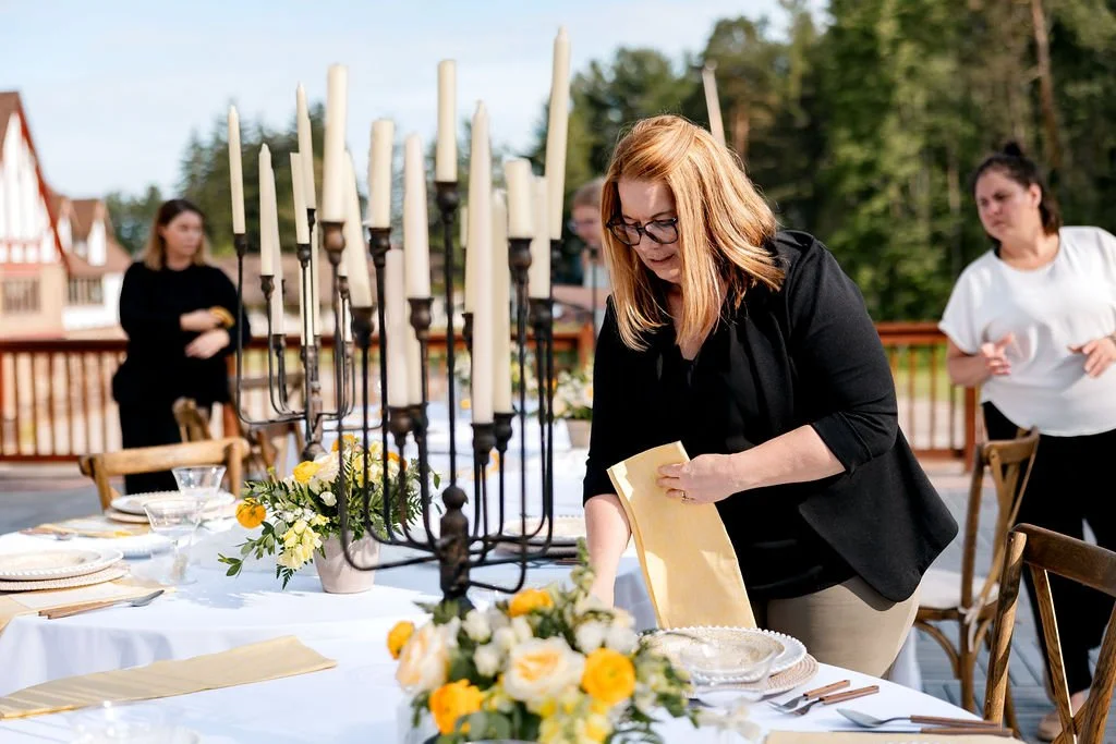 People setting up an outdoor table for a formal event with candles, flowers, and tableware.