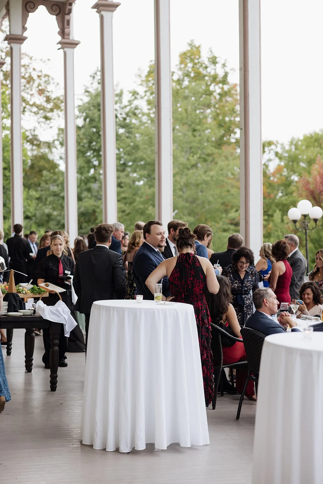 People gathering at an outdoor event or celebration, standing and sitting around high-top tables, with some serving food and drinks, in a venue with large windows and a lush green outdoor background.