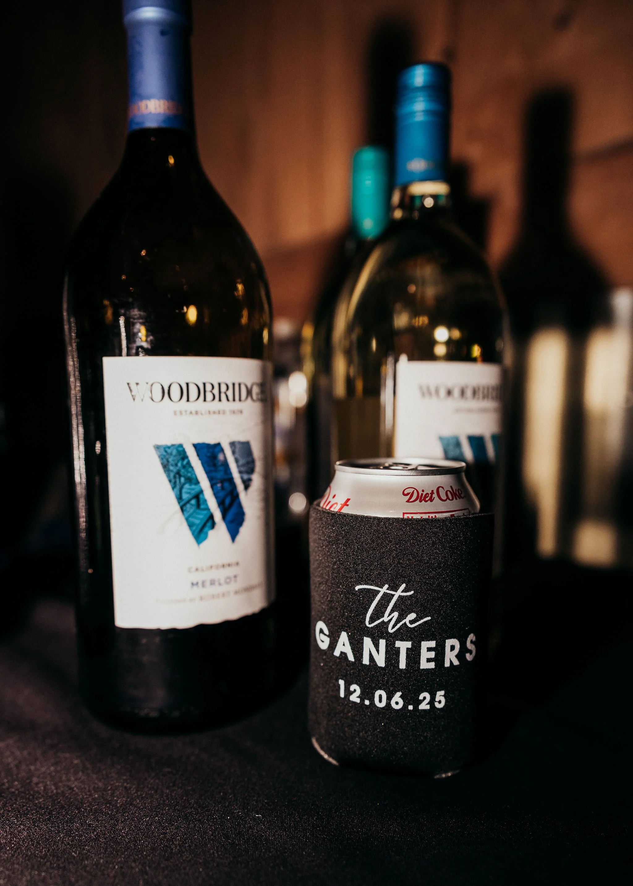 A close-up of several wine bottles and a beverage can on a dark surface. The bottle on the left is a Woodbridge Merlot with a blue and white label. In the foreground is a black can with white text and red accents, featuring a personalized message wit