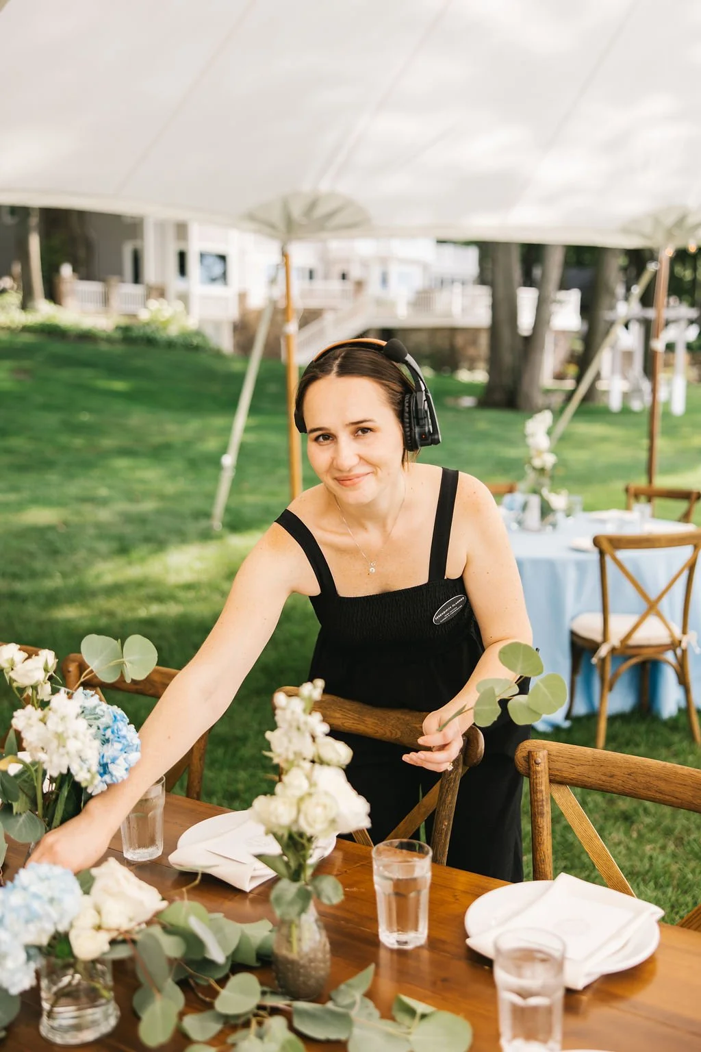 A birthday party setting outdoors with a woman wearing a black dress and a headset, arranging flowers on a wooden table under a white tent.