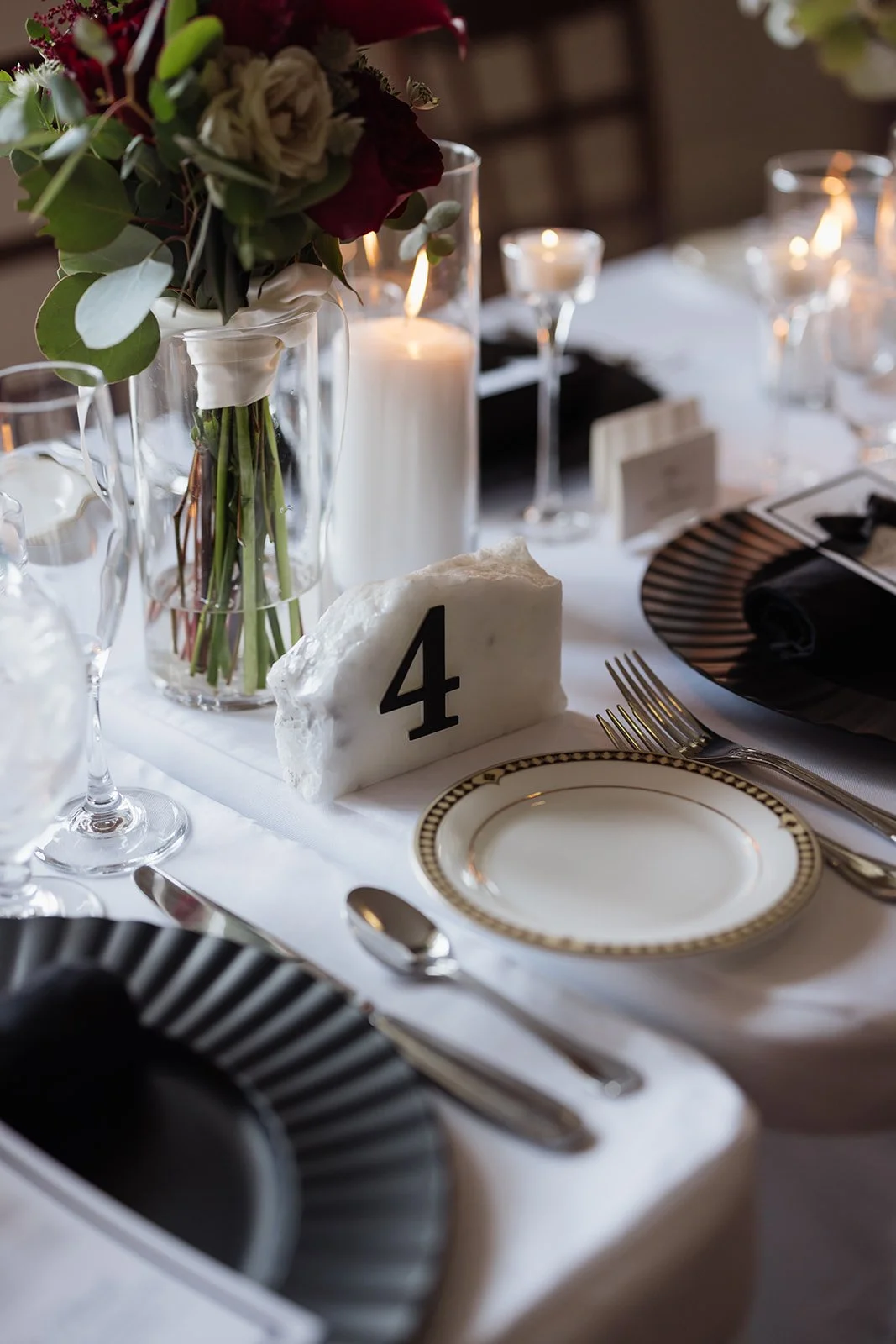 Elegant table setting with a floral centerpiece, white candles, a table number 4 in a marble holder, fine china, silverware, and black napkins, set for a formal event.