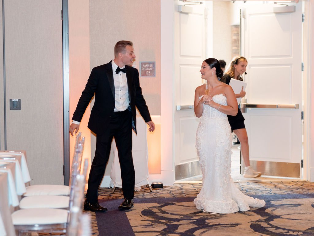 Bride and groom at their wedding reception, the groom in a tuxedo and the bride in a white wedding gown, holding a glass and smiling at each other. A woman in black is walking past in the background.