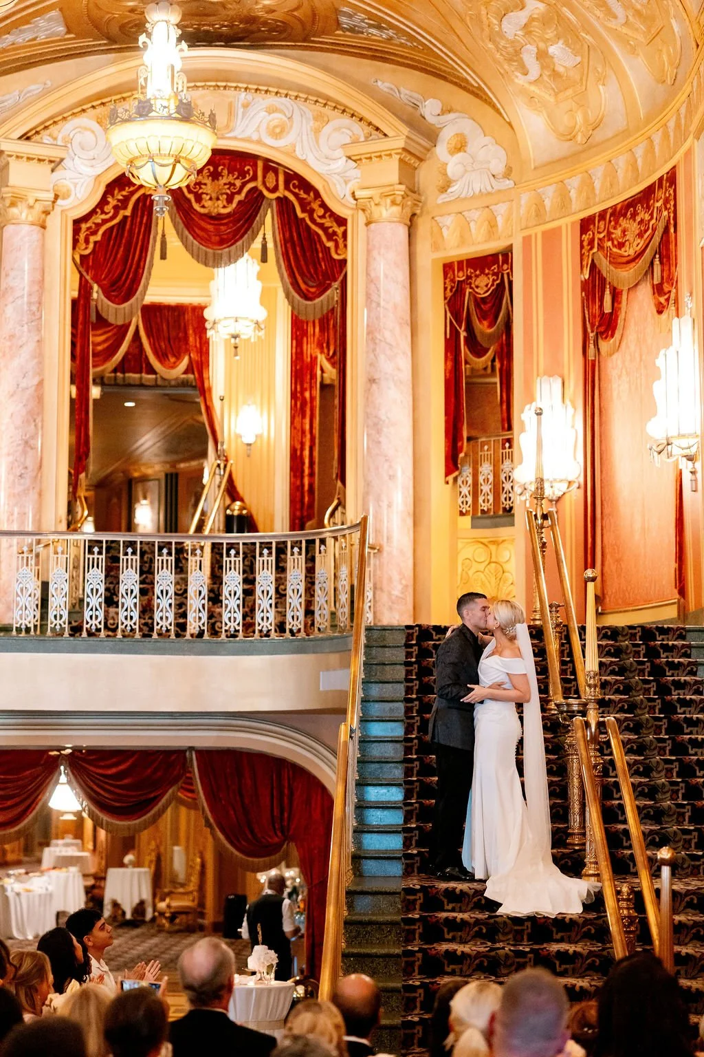 A bride and groom sharing a kiss on a staircase at their wedding reception in a grand, ornate hall with red velvet curtains, gold accents, chandeliers, and decorative ceiling moldings.