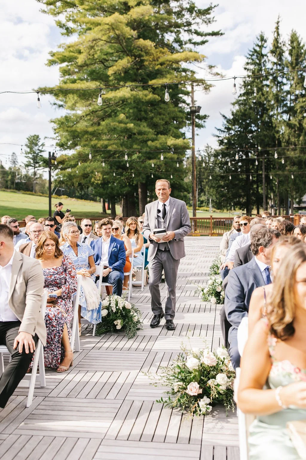 An outdoor wedding ceremony with guests seated on wooden chairs, a man in a gray suit holding a book walking down an aisle decorated with white flowers, string lights overhead, large trees in the background, and a partly cloudy sky.