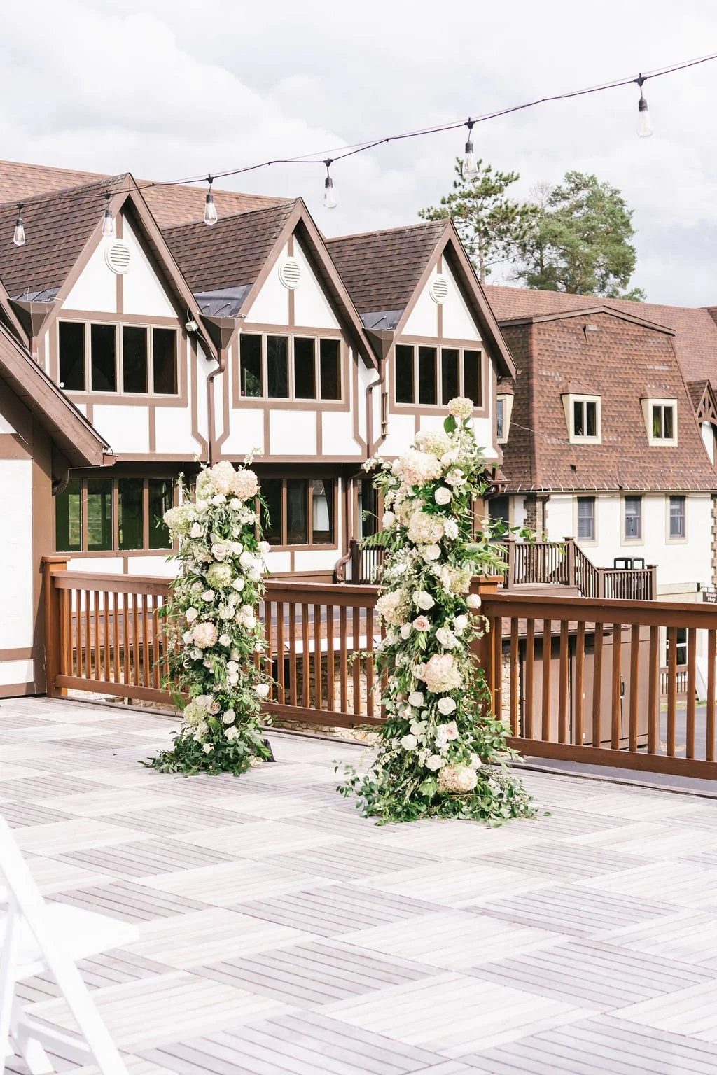 Outdoor wedding setup on a terrace with floral decorations, in front of a Tudor-style building with brown shingled roof and large windows, overcast sky.