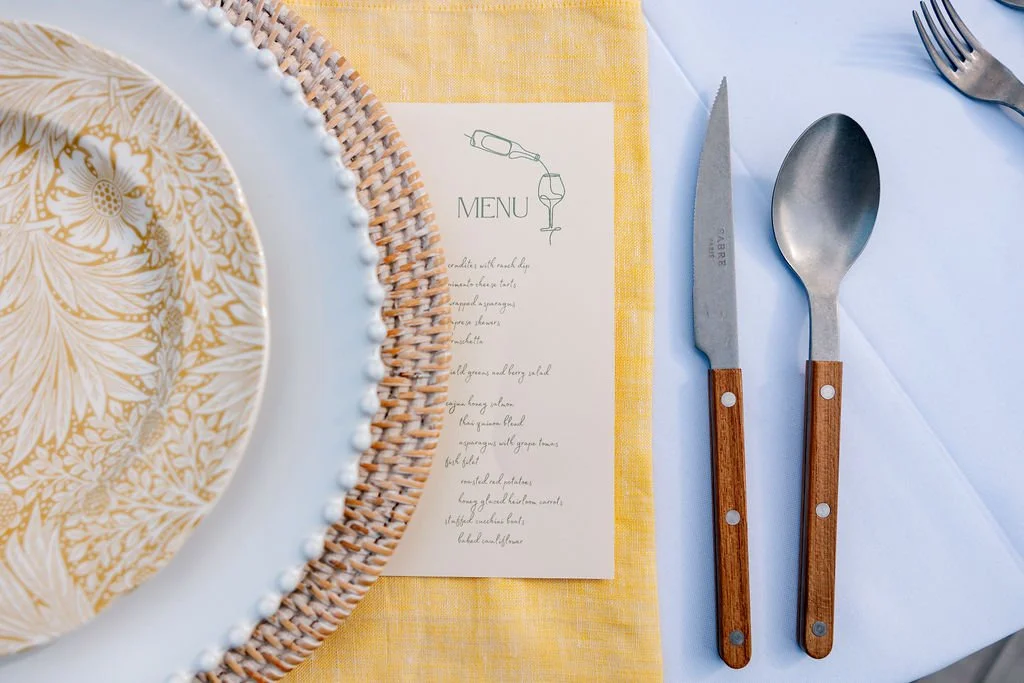 A neatly arranged table setting with a decorative plate, a menu card, and cutlery including a knife and spoon with wooden handles on a white and yellow tablecloth.