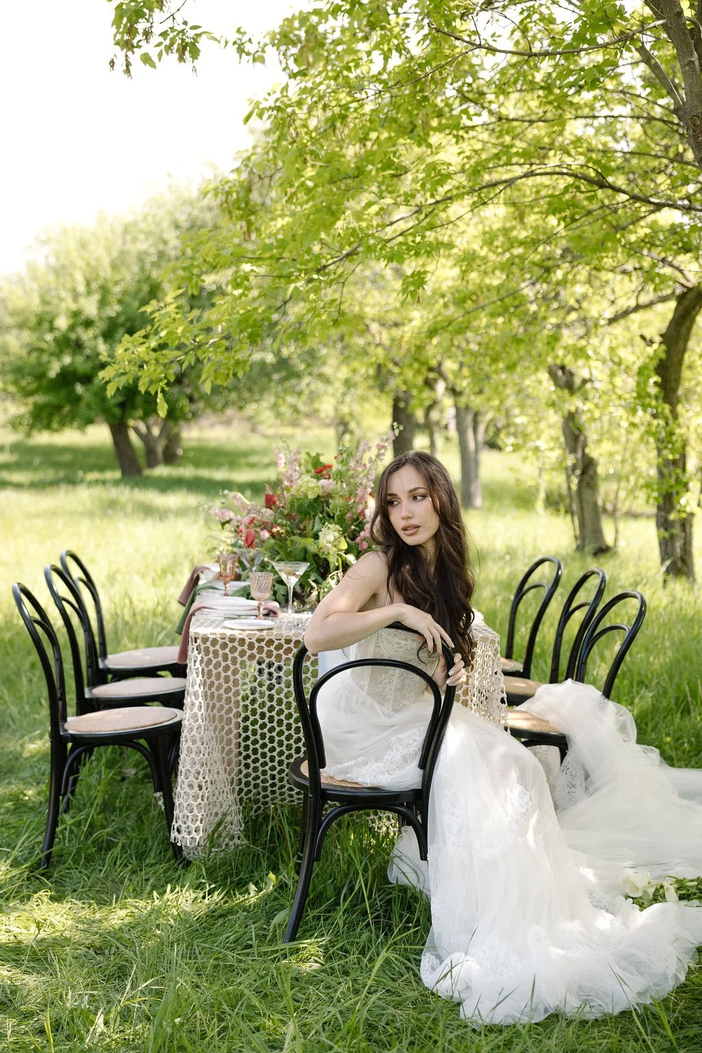 A woman in a white dress sitting at a decorated table outdoors in a lush green garden with trees.