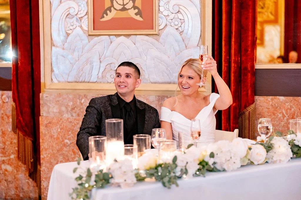 A bride and groom seated at a wedding reception table, smiling. The bride holds a glass of champagne raised in a toast, and the table is decorated with white flowers and candles.