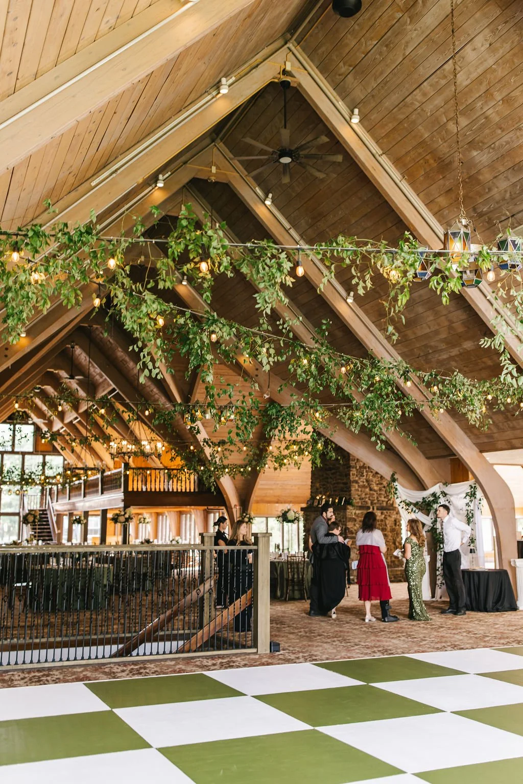 People at a celebration or wedding reception in a rustic barn with wooden beams, greenery, string lights, and a checkered dance floor.