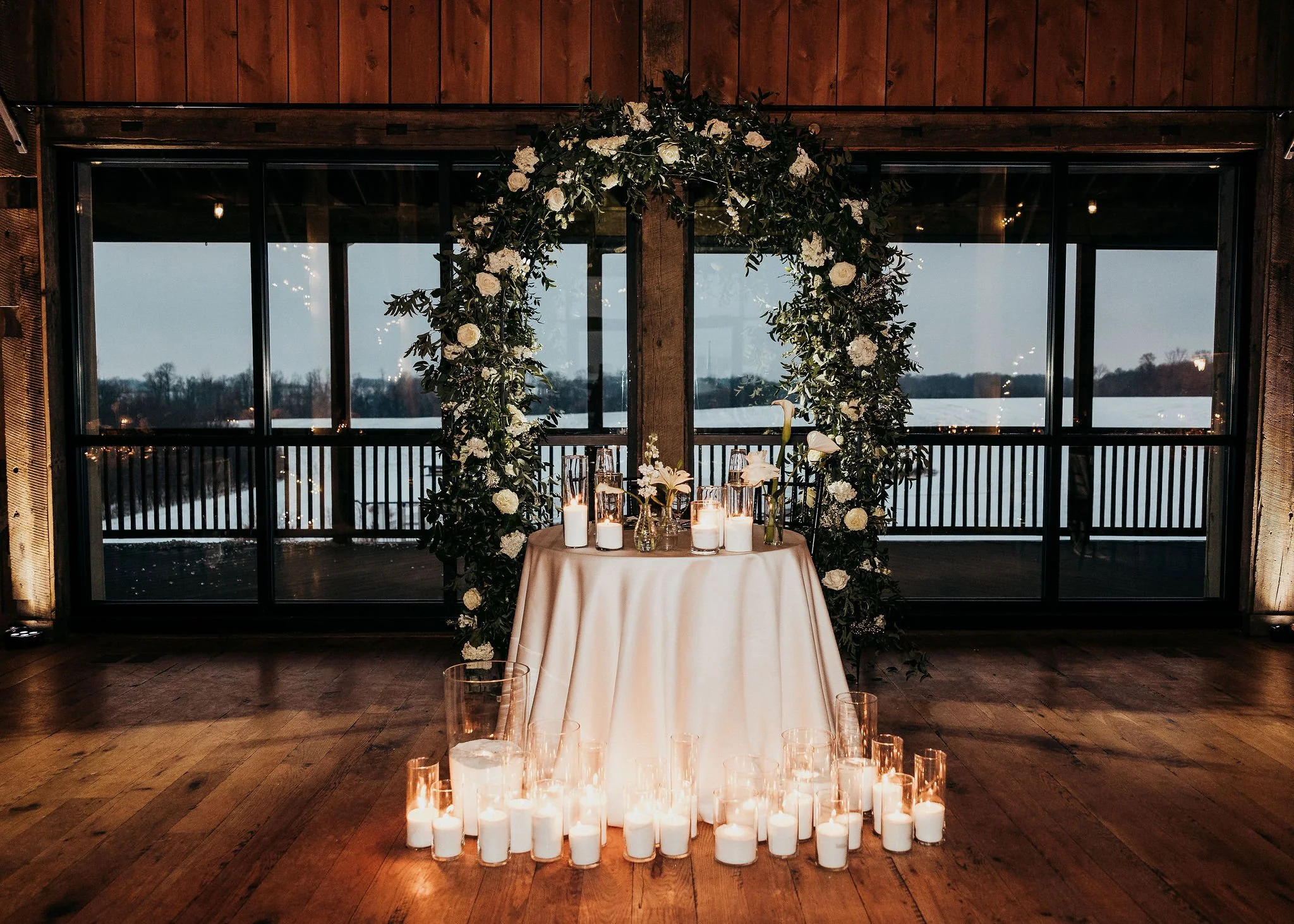 A round table with a beige tablecloth, decorated with white flowers in vases, surrounded by candles in glass holders, set in front of large windows with a winter landscape outside. A large floral arch decorated with white roses and greenery is behind