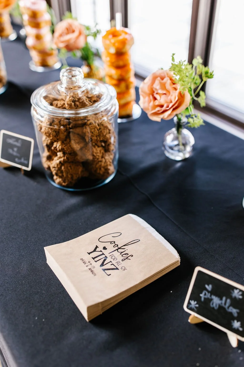 Close-up of a table with a glass jar filled with cookies, a small vase with peach-colored flowers, and a stack of brown paper napkins that reads 'Cookies for all of us ZINK RAN & AURORA'.