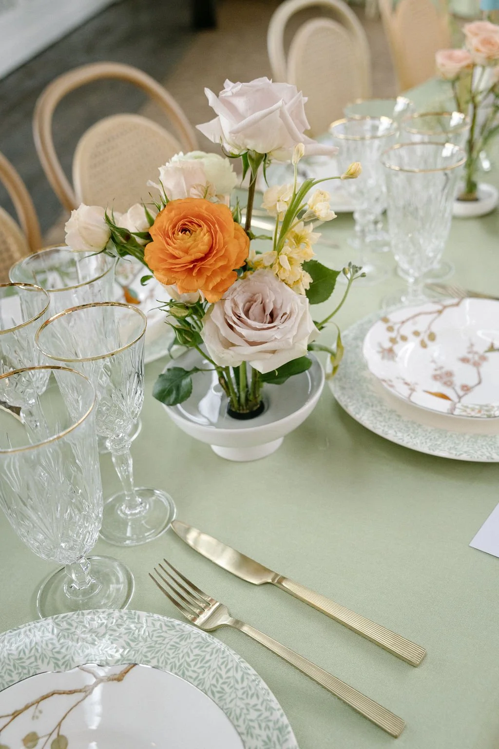 Elegant table setting with a floral centerpiece featuring white, orange, and pale pink roses and yellow blossoms, surrounded by crystal glasses, gold-rimmed plates, and gold flatware on a light green tablecloth.