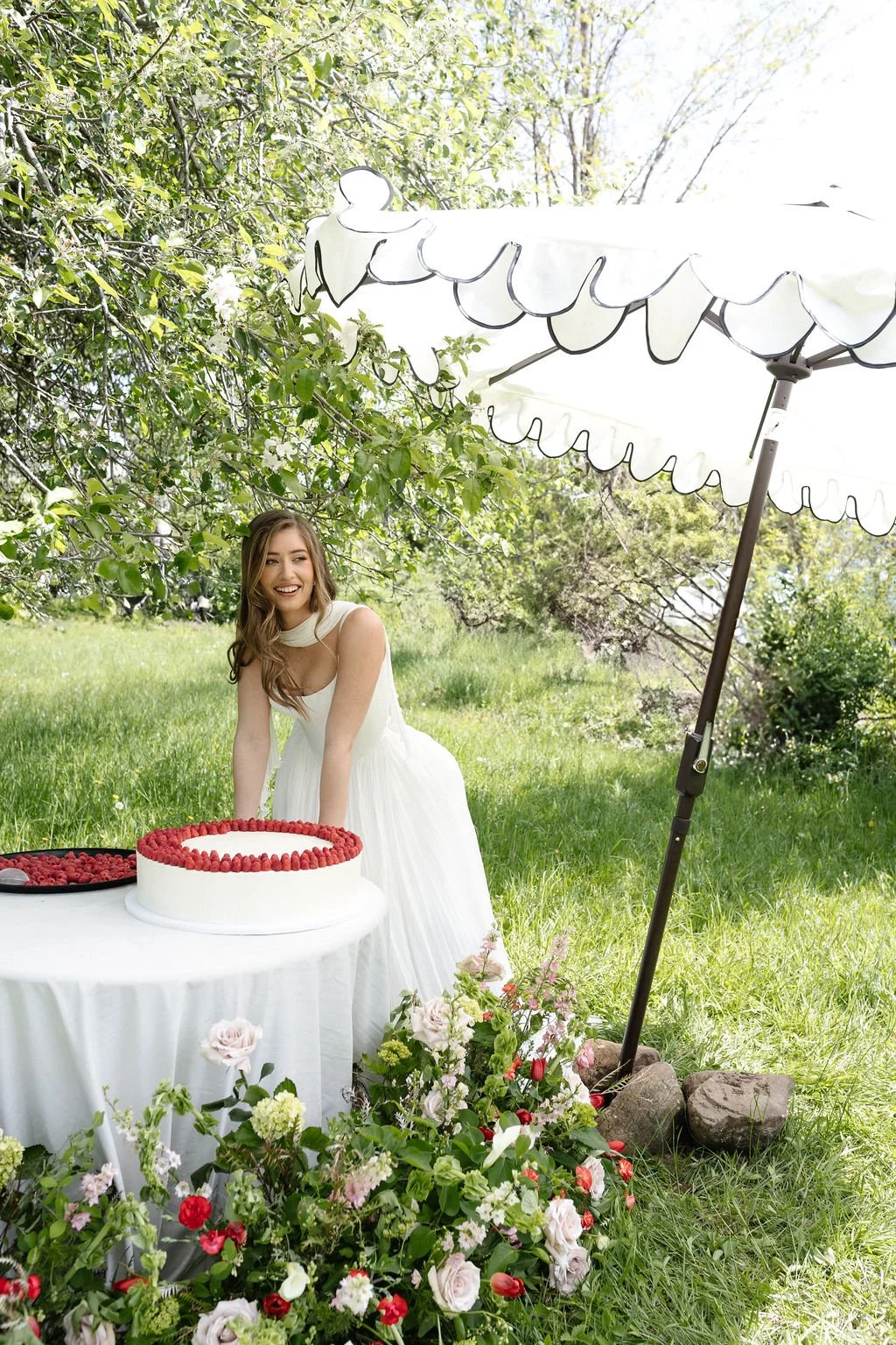A young woman in a white dress smiling and celebrating outdoors under a large white umbrella, with a cake decorated with raspberries on a table, surrounded by flowers and greenery.