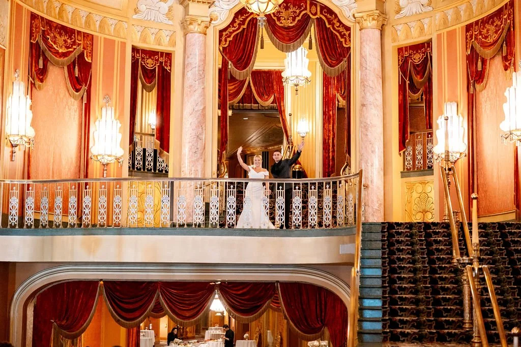 A bride and groom standing on a balcony inside a grand, luxurious venue with ornate curtains, chandeliers, and intricate railings, celebrating their wedding.