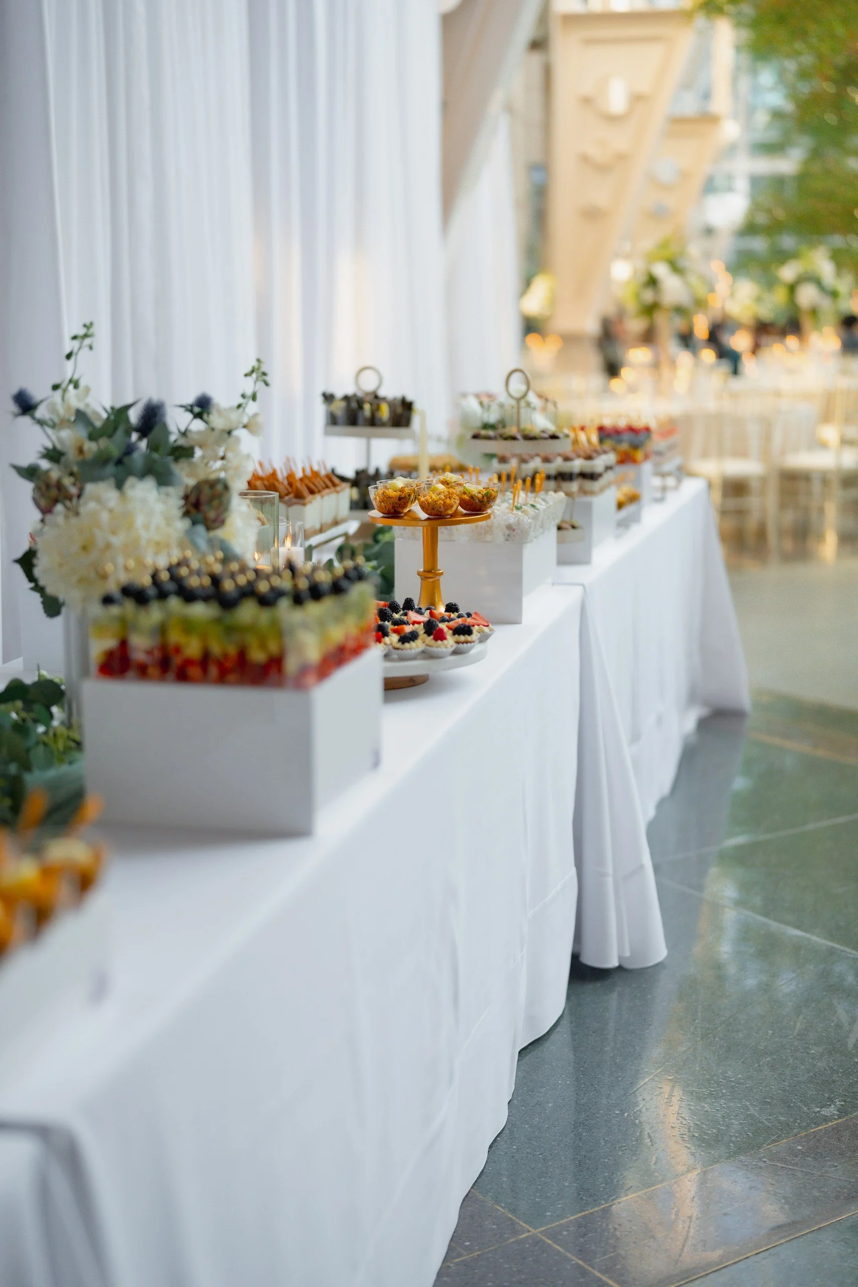 A table with various desserts and floral arrangements at what appears to be a wedding or event reception, with a bright and elegant setting.