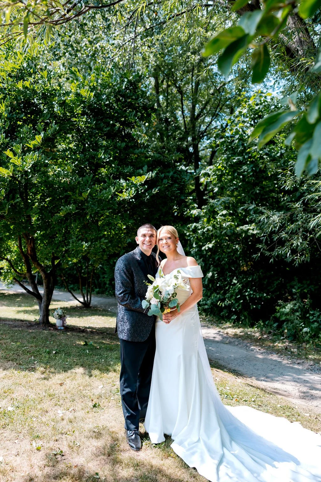 Bride and groom standing outdoors on a sunny day, surrounded by green trees, smiling, with the bride holding a bouquet of white flowers and greenery.