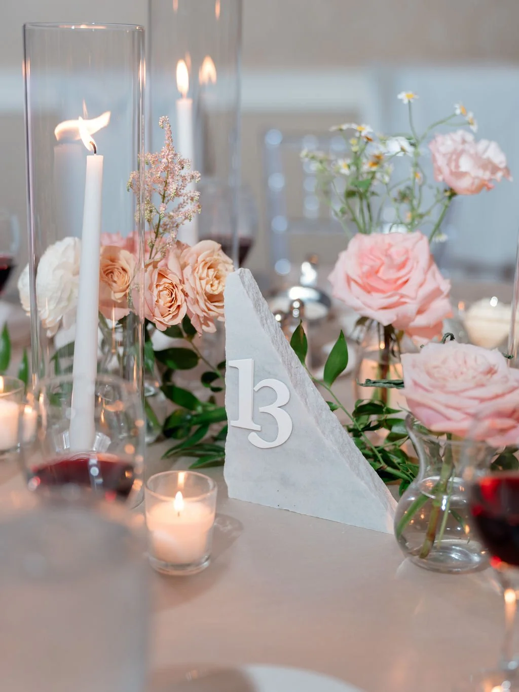 Elegant table centerpiece with pink roses, small white flowers, candles, and a table number 13, set on a white tablecloth for a formal event or wedding.