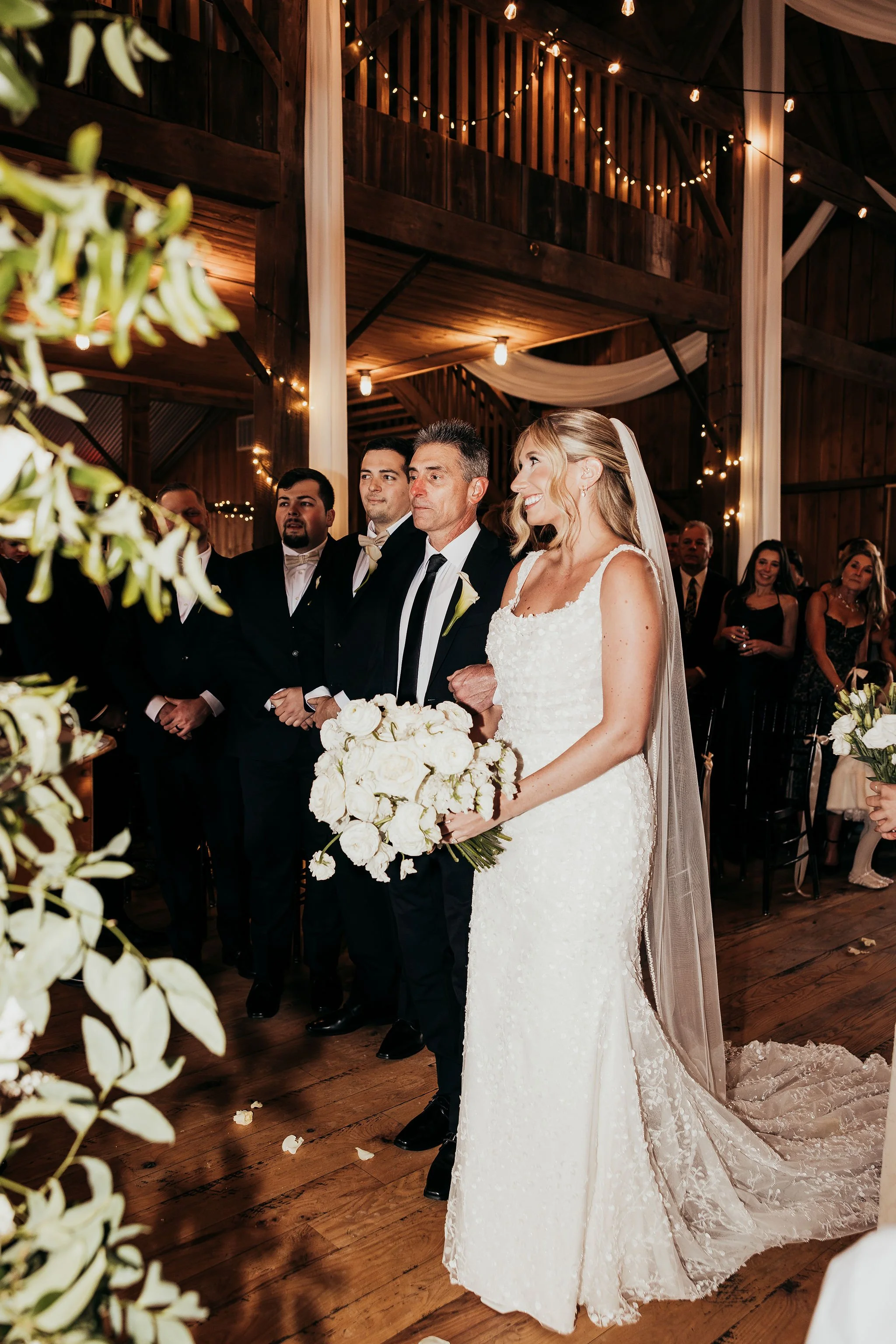 A bride and groom standing together during their wedding ceremony in a rustic barn decorated with string lights and white drapery, with guests present in the background.