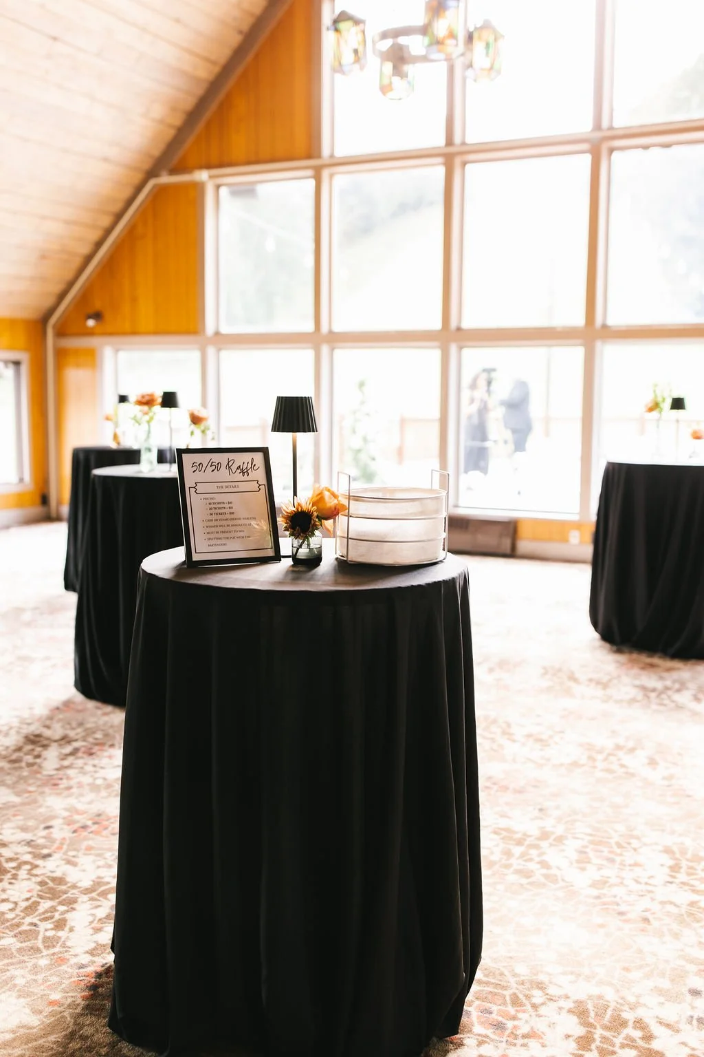 Indoor event space with round tables covered in black tablecloths, decorated with small floral arrangements and a sign for a '50/50 Raffle.' Large floor-to-ceiling windows let in natural light. The room has a wooden ceiling and walls, with colorful h