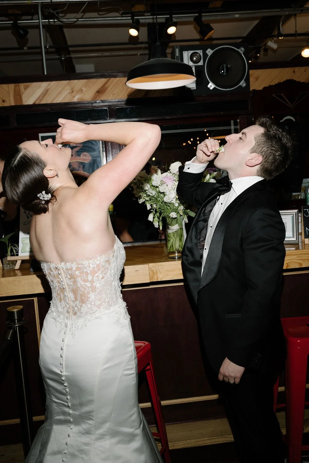 Bride and groom dressed in wedding attire, eating cake at a wedding reception in a decorated venue.
